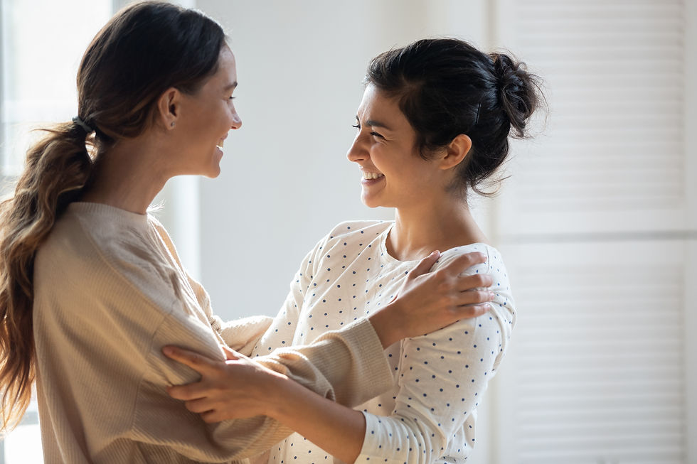 Two happy young women holding each other’s shoulders, about to hug indoors.